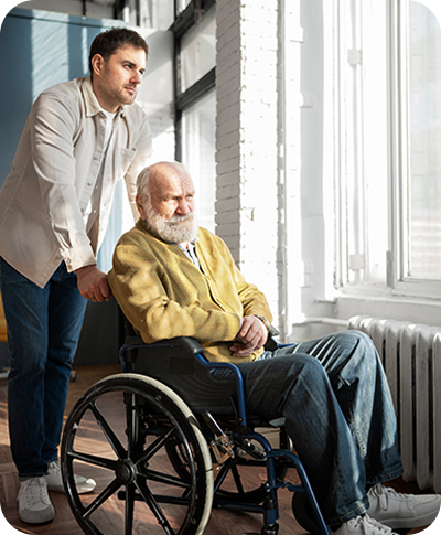 Support worker standing behind an older man in a wheelchair beside a sunlit window